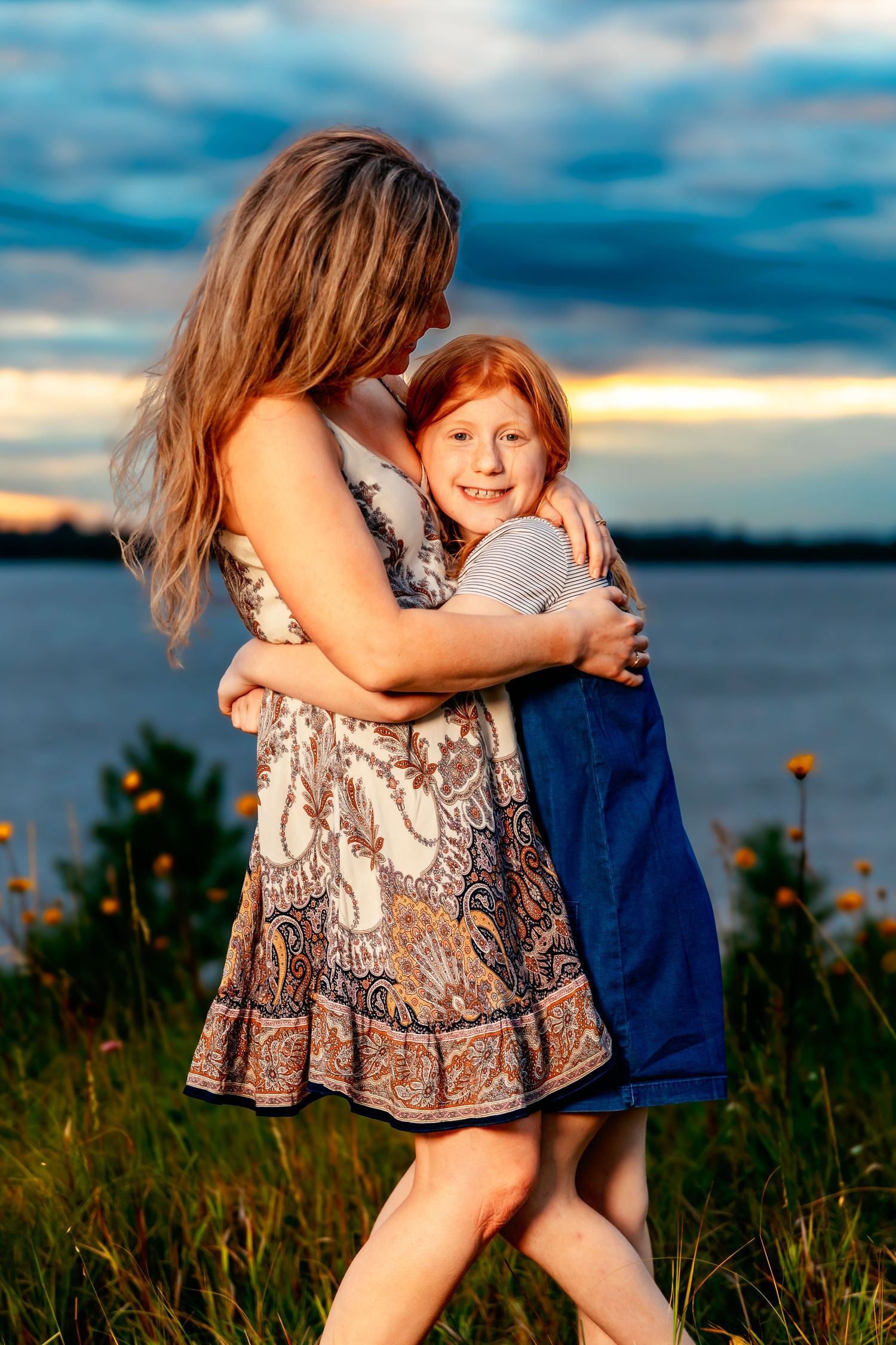 Mother and daughter share a tender embrace at sunset by a lake with wildflowers in the foreground.