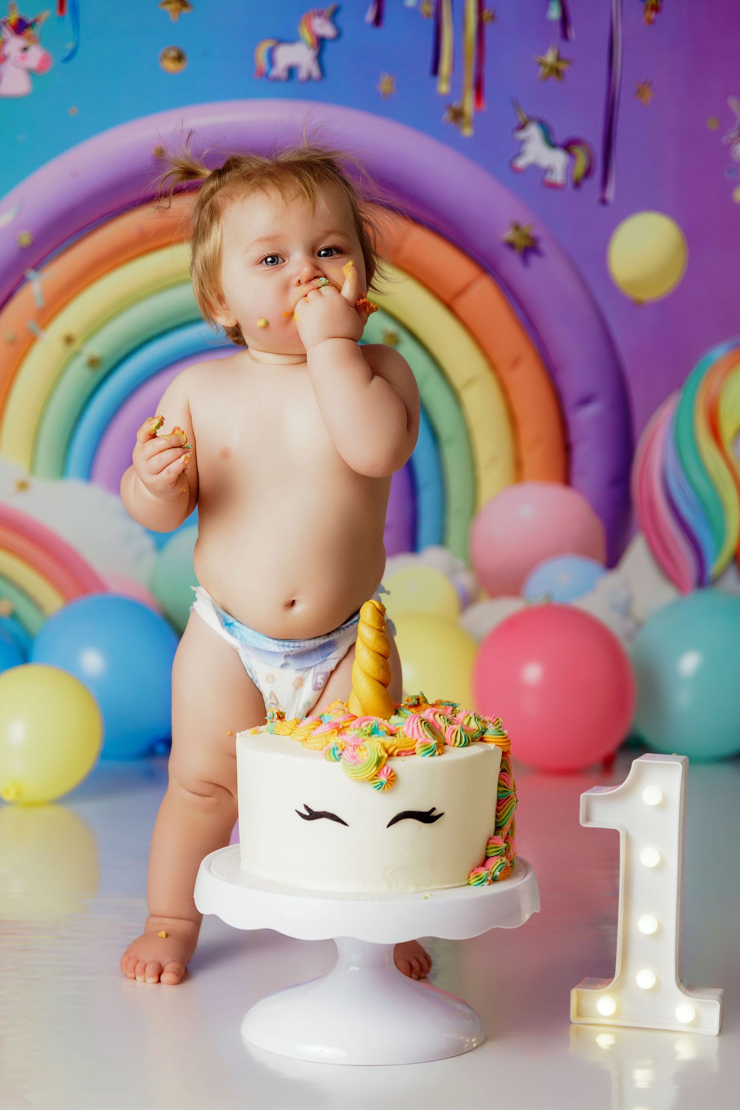 Baby in diaper smashing unicorn cake at colorful rainbow first birthday party photo shoot.
