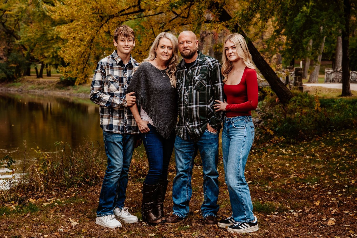 A family of four poses together on an autumn day by a pond with golden fall foliage in the background.