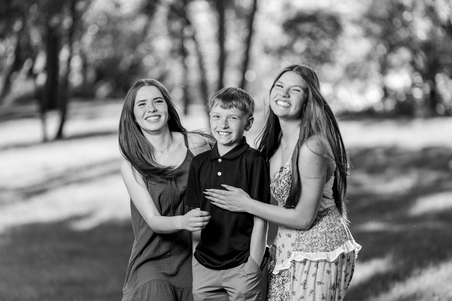 Black and white photo of two teenage girls and a young boy smiling together outdoors in a sunny park.