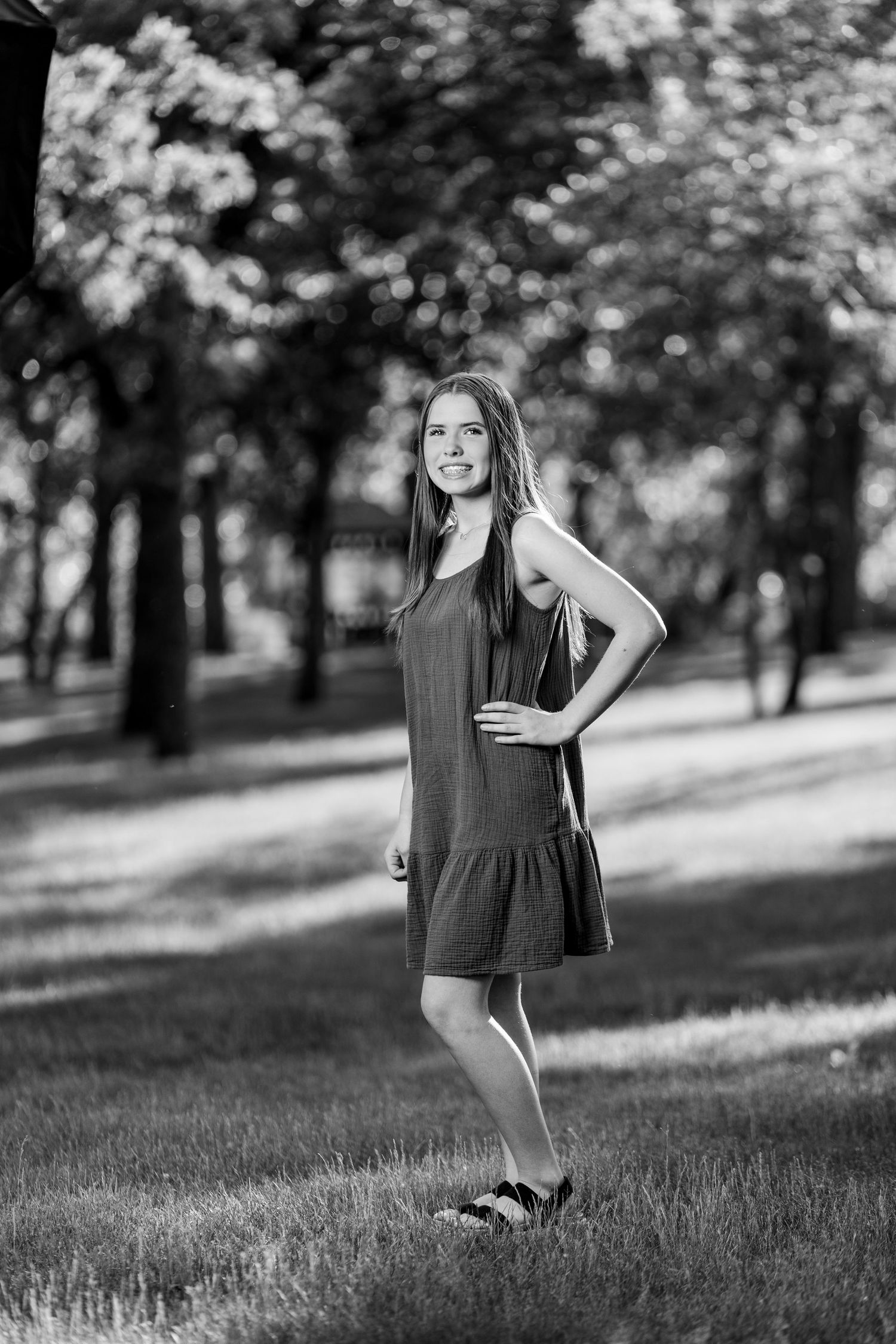 Young teenage girl in a gray dress posing in a tree-lined park in a black and white photo.