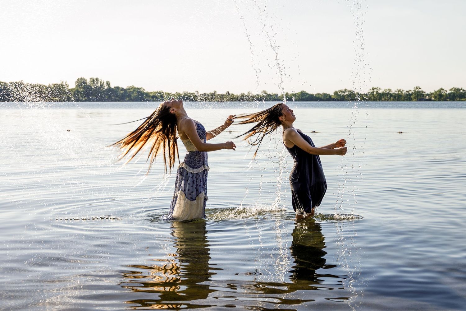 Two girls tossing their wet hair while standing in a lake on a sunny day.