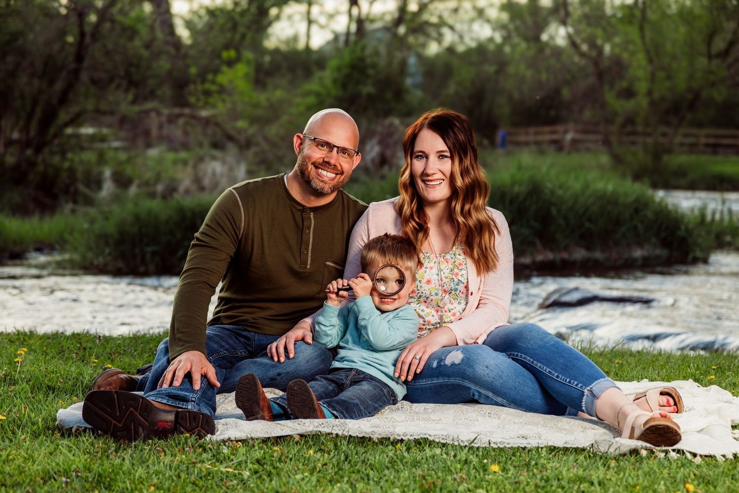 Happy family of three smiling together on a blanket by a peaceful river in a lush green park.