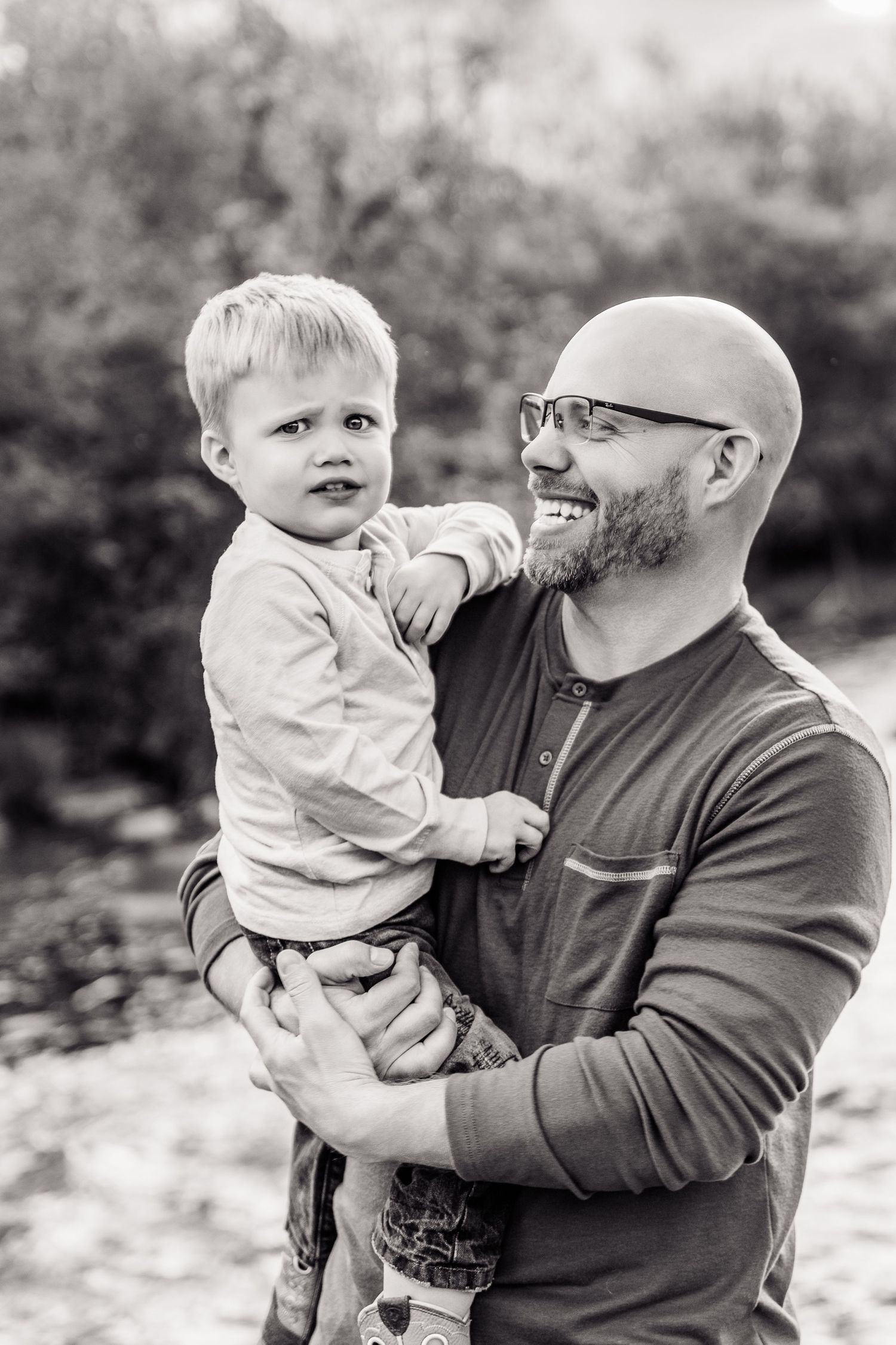 Black and white photo of a bald bearded father smiling while holding his young blonde toddler son outdoors.