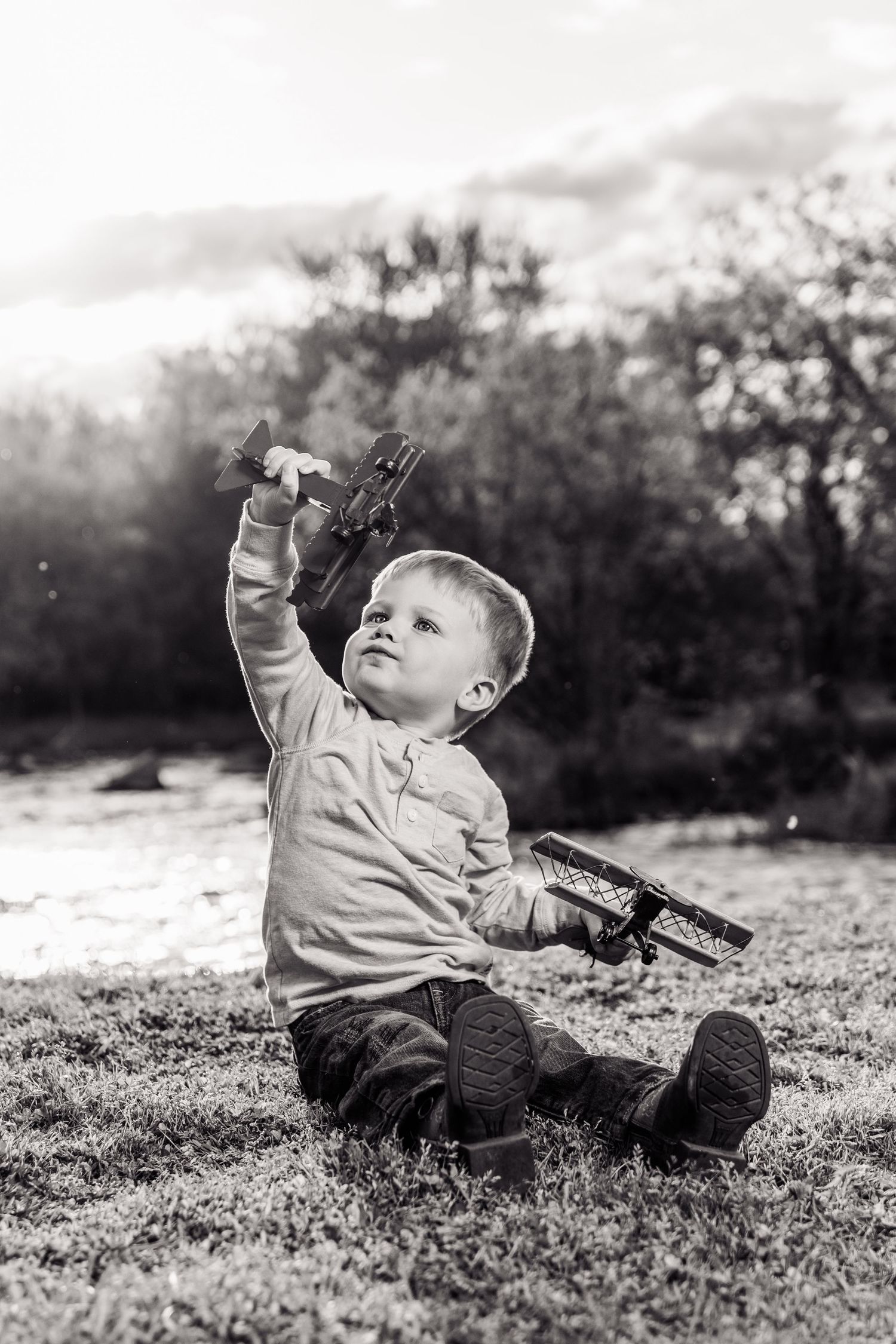 Black and white photo of a young boy sitting on grass, holding a toy airplane up in the air outdoors.