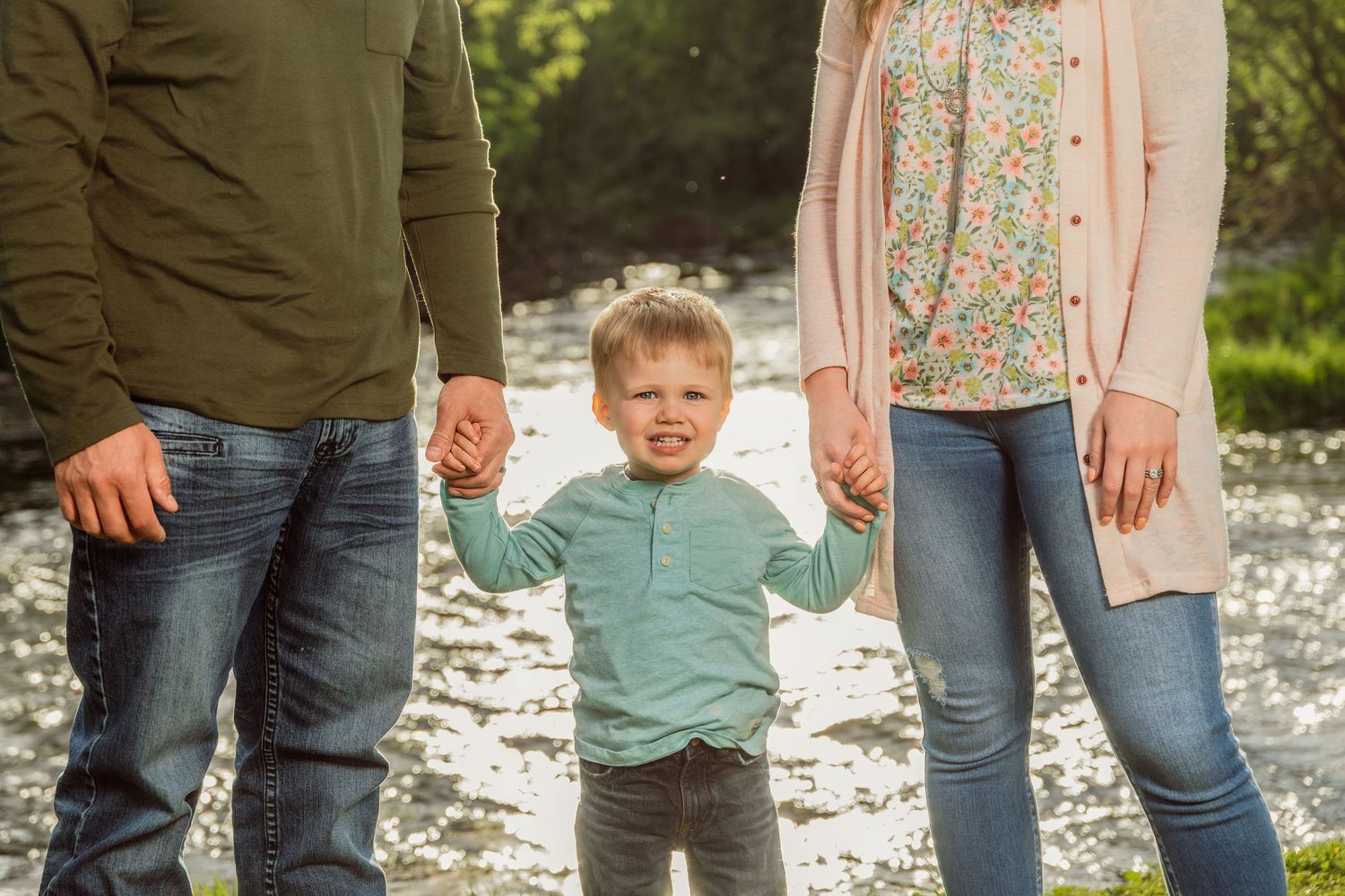 Young boy holds hands with parents outdoors near a stream, wearing a teal shirt in a sunny nature setting.