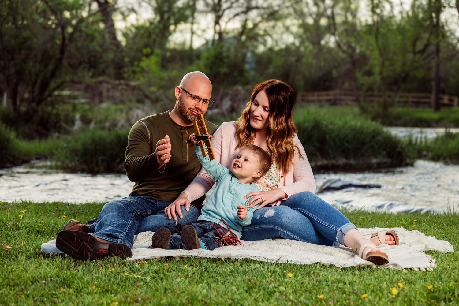 Family of three enjoying a picnic on a blanket by a pond in a lush green park setting.