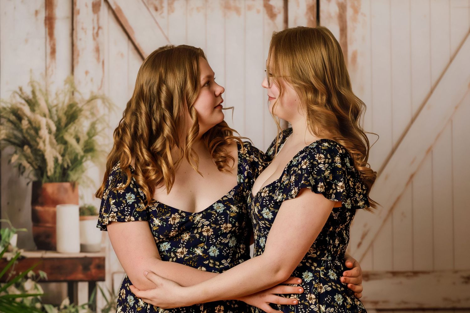 Two women in matching floral dresses embrace and smile at each other in a rustic studio setting.