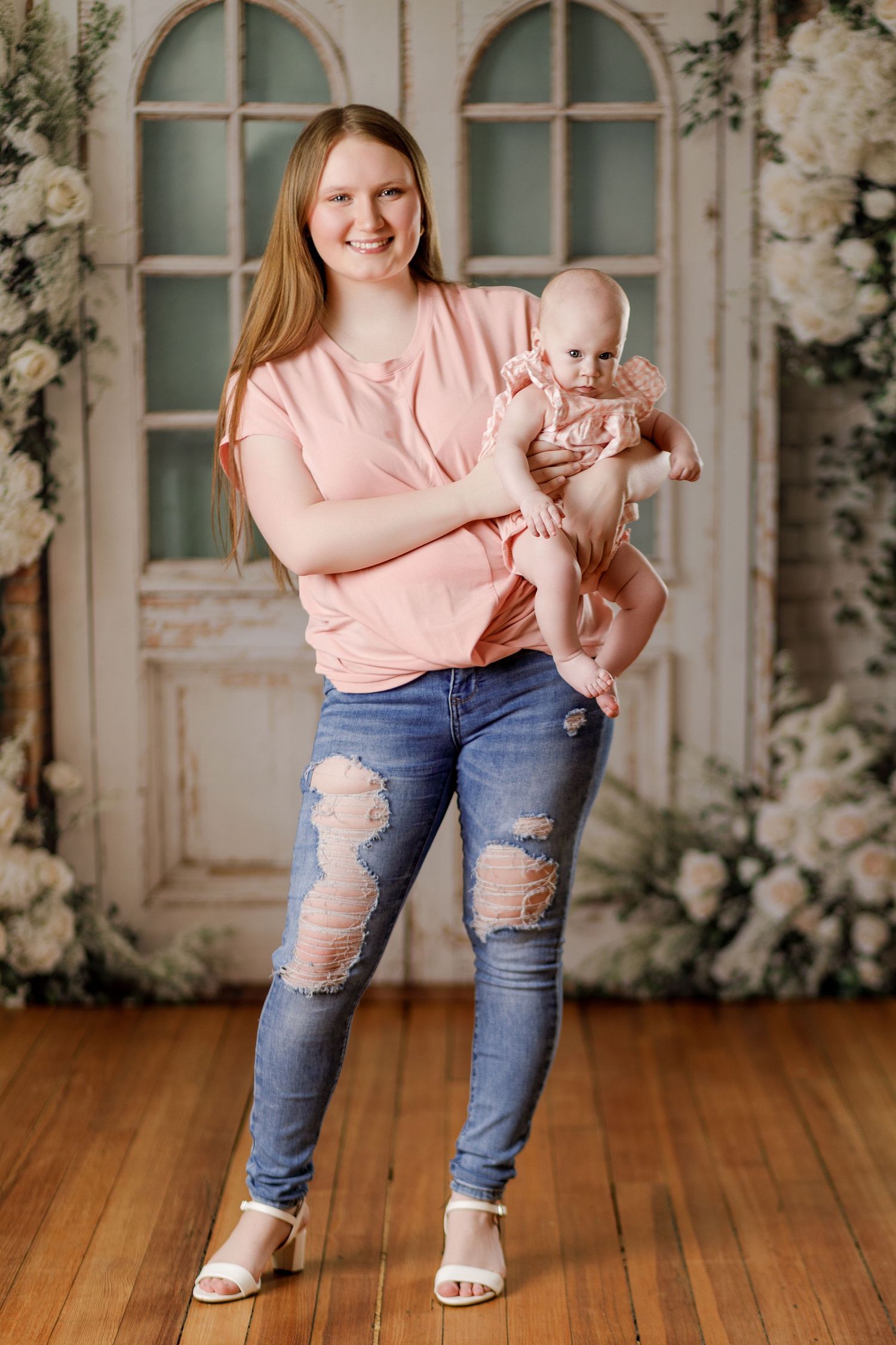 Young mother in pink top and ripped jeans holds smiling baby in floral studio portrait.