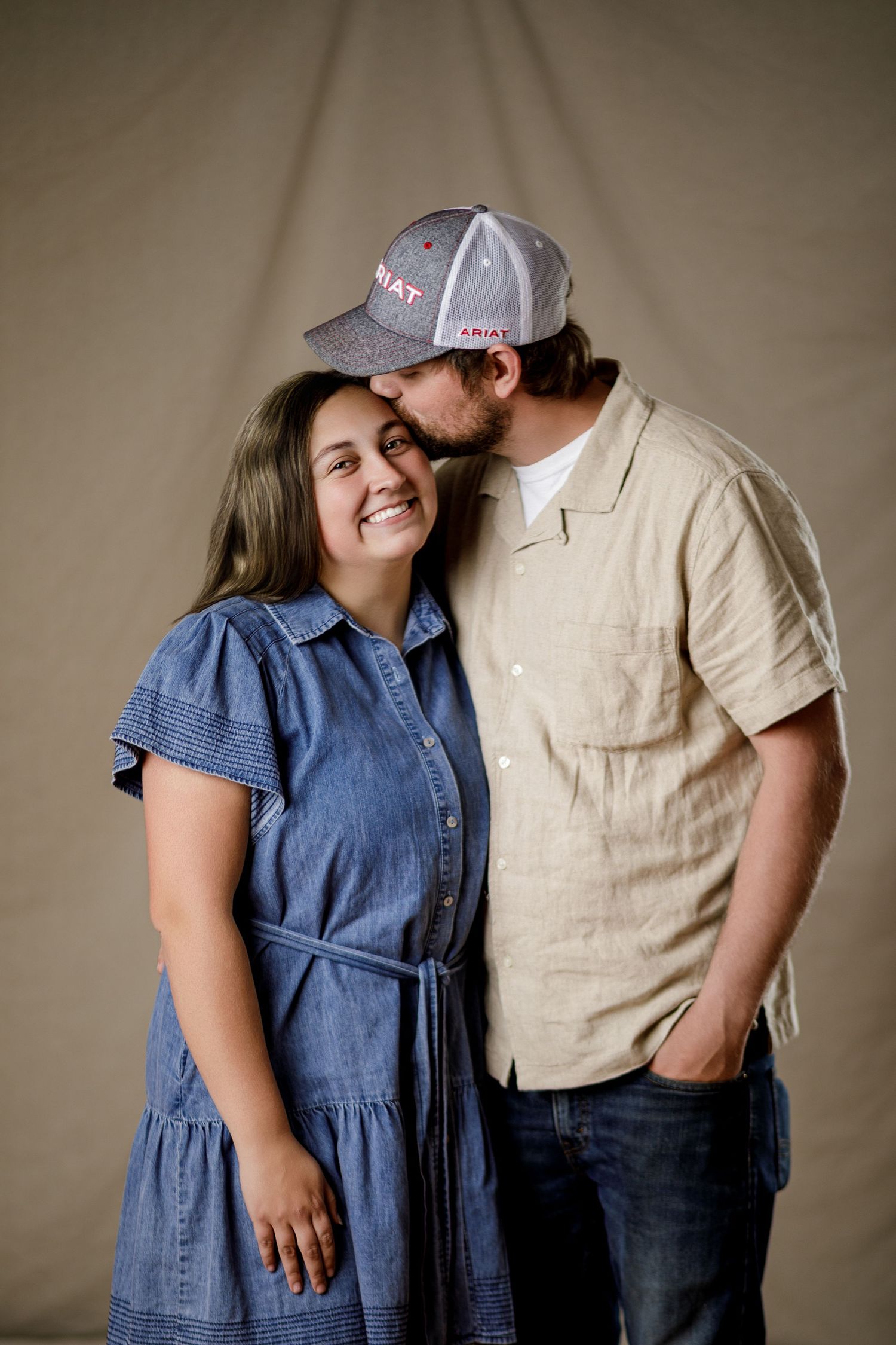 Couple posing together, man in cap kissing woman's head as she smiles in blue denim dress.