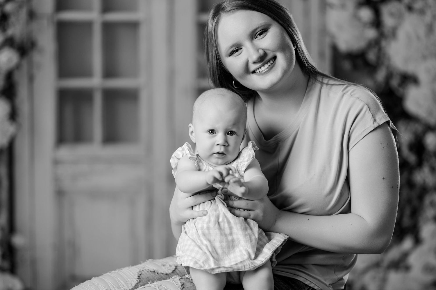 Black and white portrait of a smiling mother holding her baby girl in a dress during a studio session.