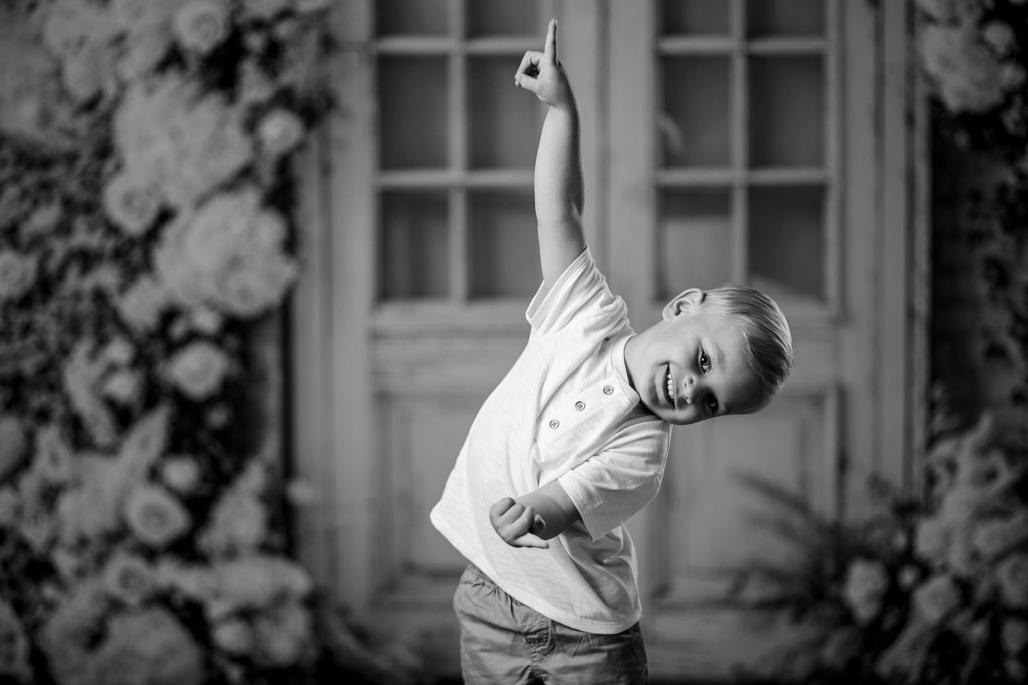 Black and white photo of a joyful child striking a fun pose with arm raised, surrounded by floral backdrop.
