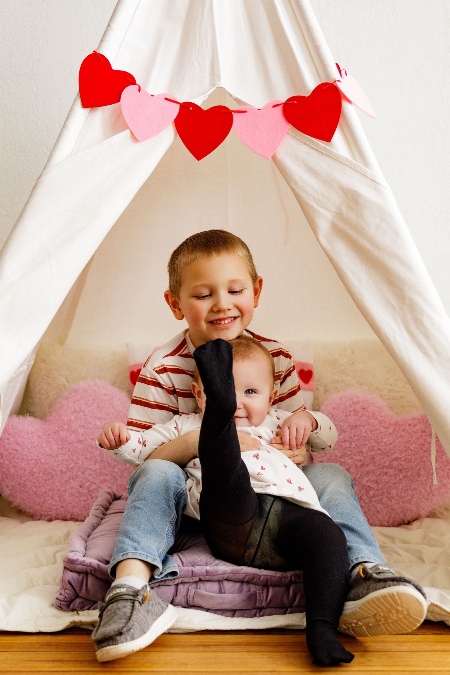 Children play and laugh together in a cozy white tent decorated with red and pink heart garlands.