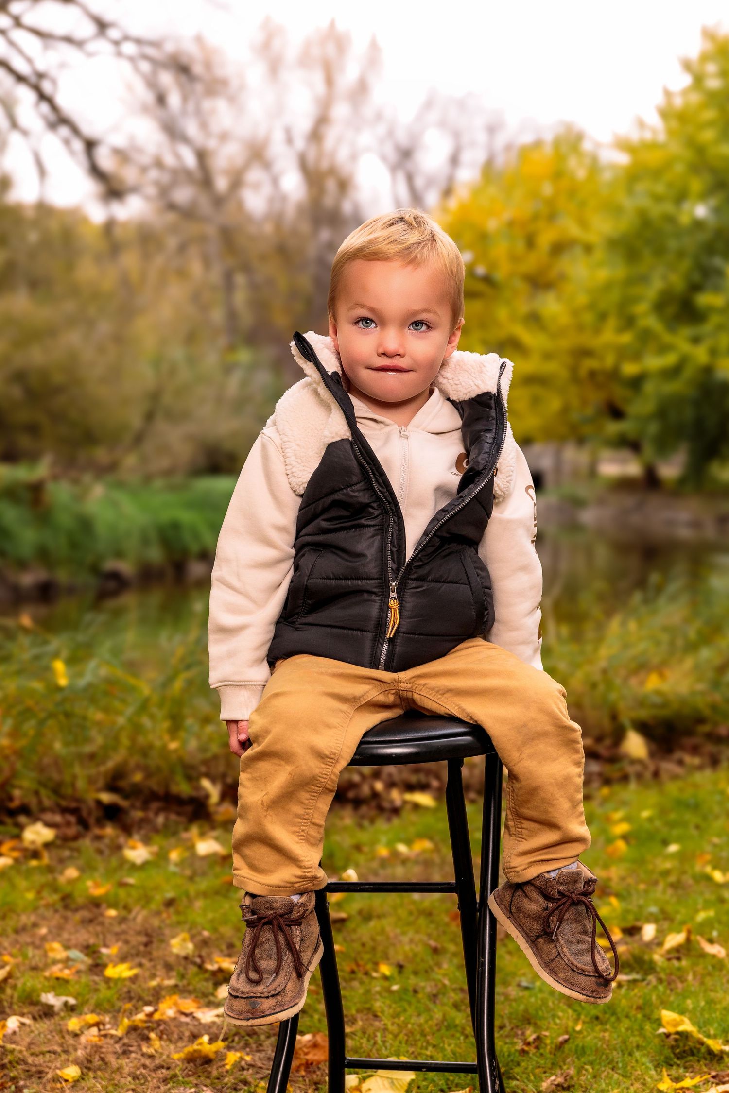 Small child in autumn attire sitting on a black chair outdoors surrounded by fallen leaves.