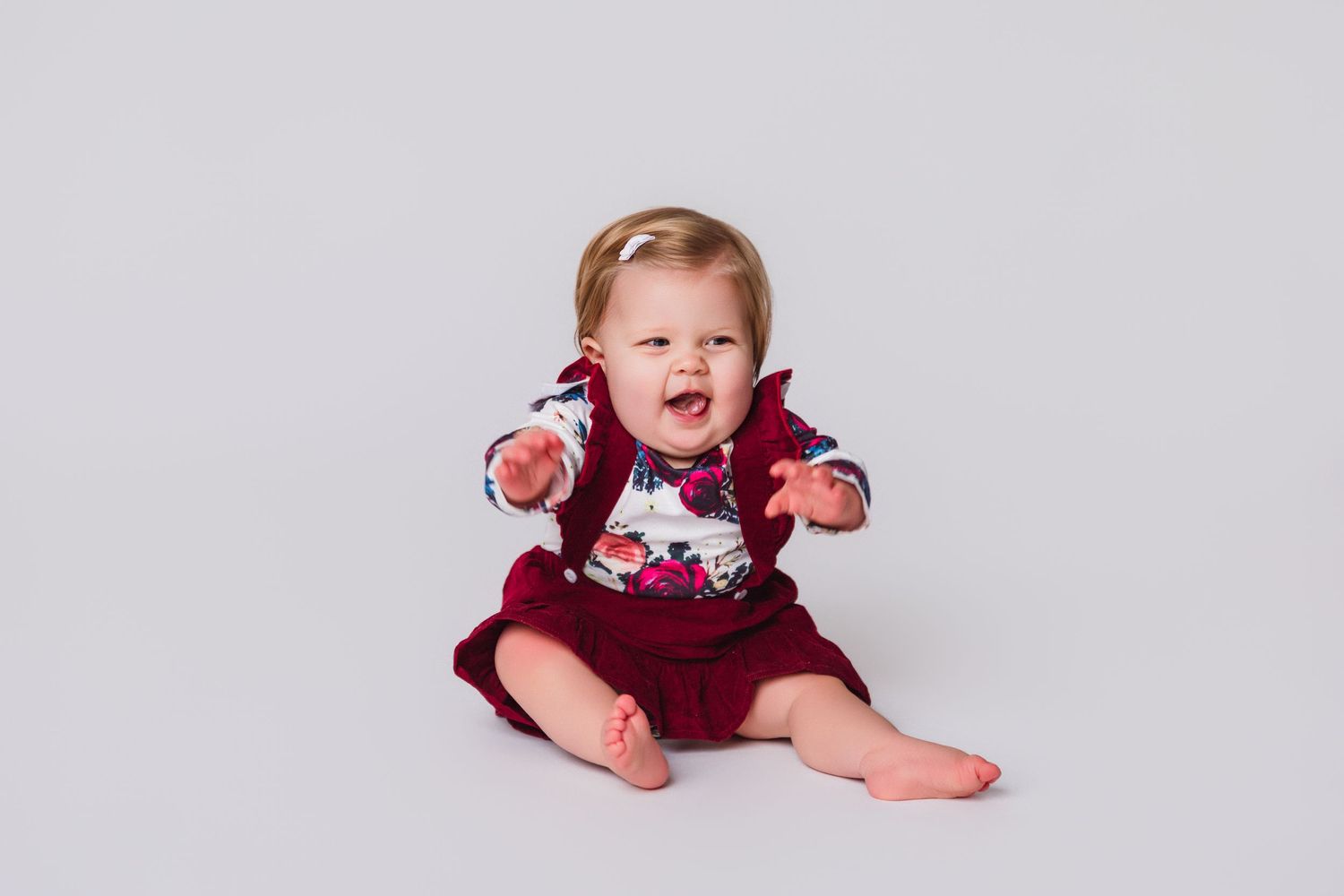 Happy baby in burgundy outfit sitting on floor against grey background.
