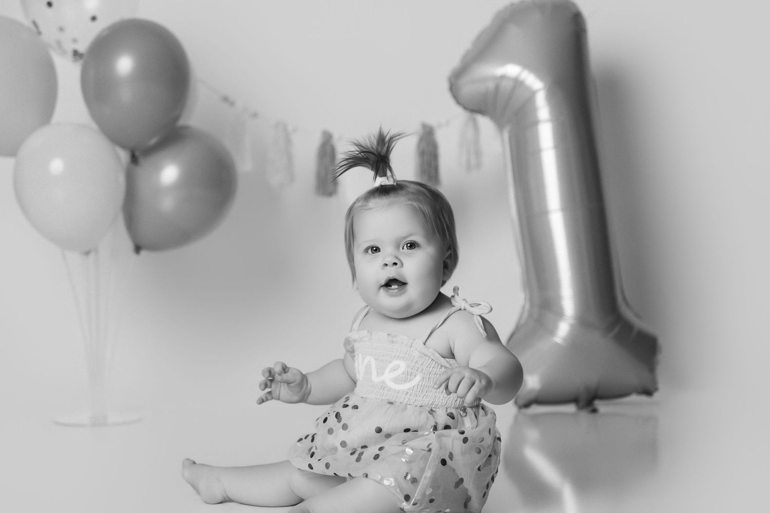 Baby wearing floral dress with number one balloon and party decorations for first birthday.