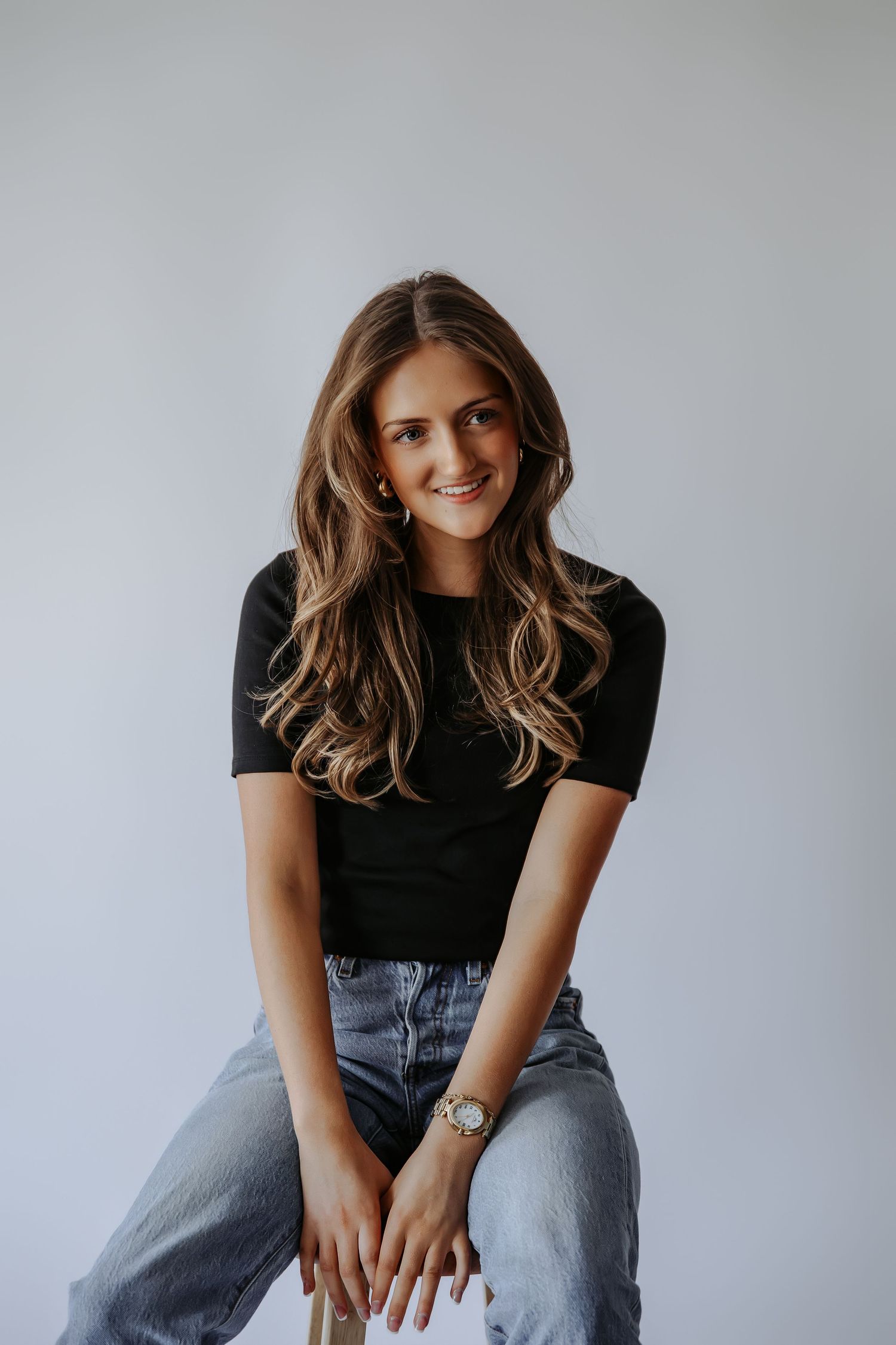 Smiling brunette woman in black t-shirt and jeans poses on stool in bright studio setting.