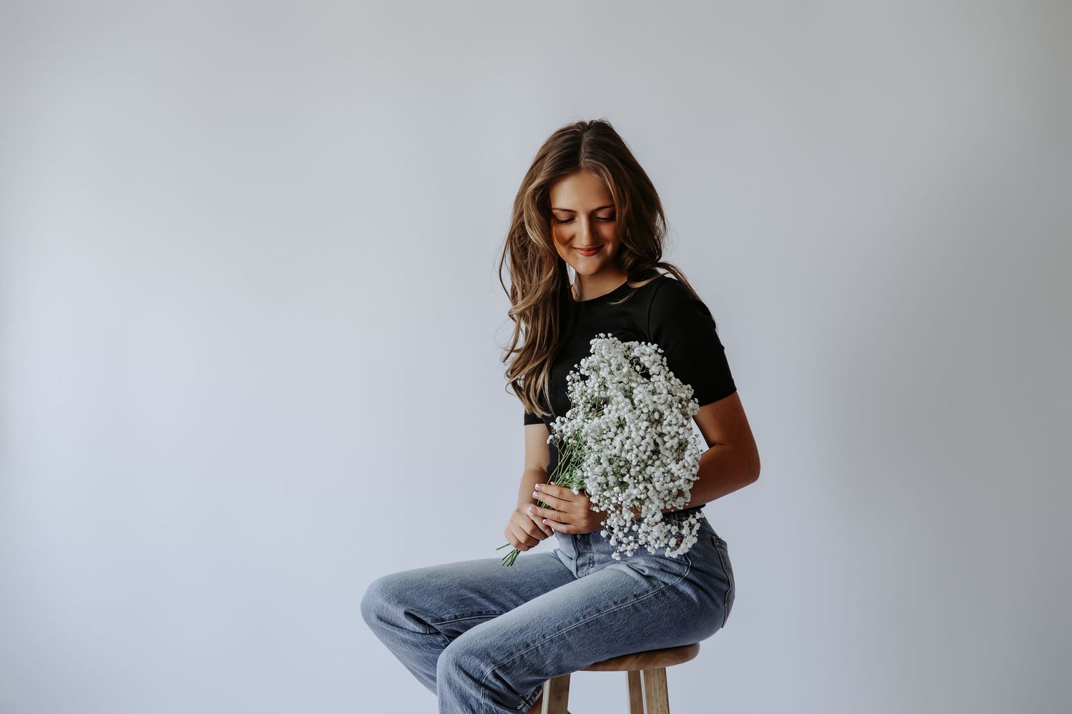 Woman in black t-shirt and jeans sitting on stool holding white baby's breath flowers, smiling downward.