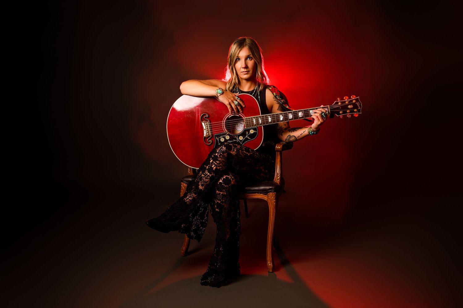 Female guitarist sits on chair holding red acoustic guitar, wearing black outfit, against dramatic red-lit dark studio background.