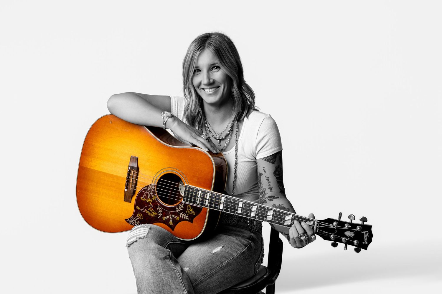 Smiling tattooed female musician holding sunburst acoustic guitar in black and white studio photo.