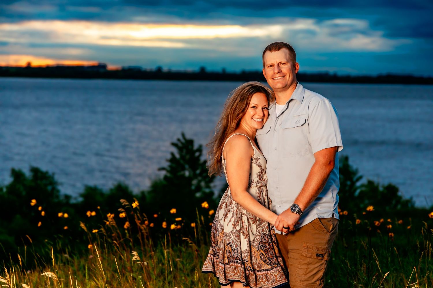 A couple shares an intimate moment during a golden hour photoshoot near a lake with tall grass and dramatic clouds.