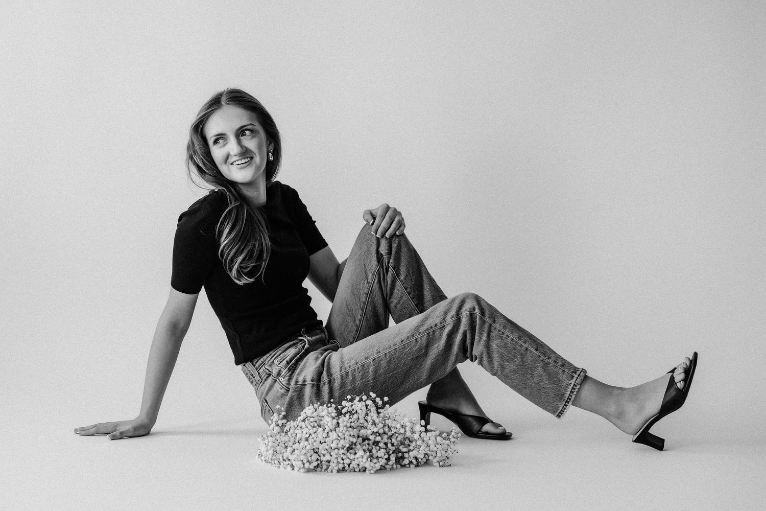 Black and white photo of a smiling woman sitting on floor in jeans and black top with flowers nearby.