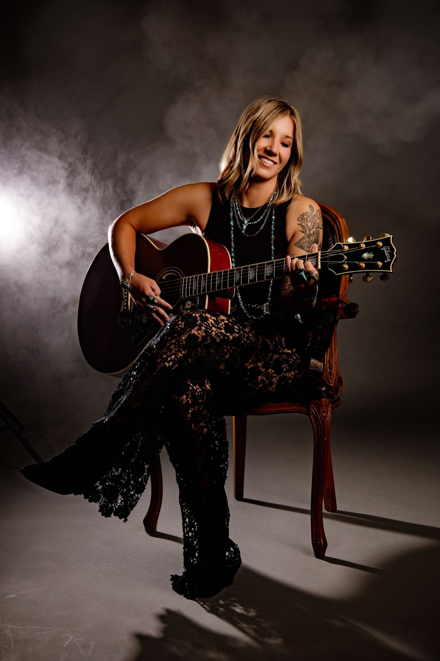 Smiling tattooed woman in lace outfit playing acoustic guitar seated in ornate chair with smoky backdrop.