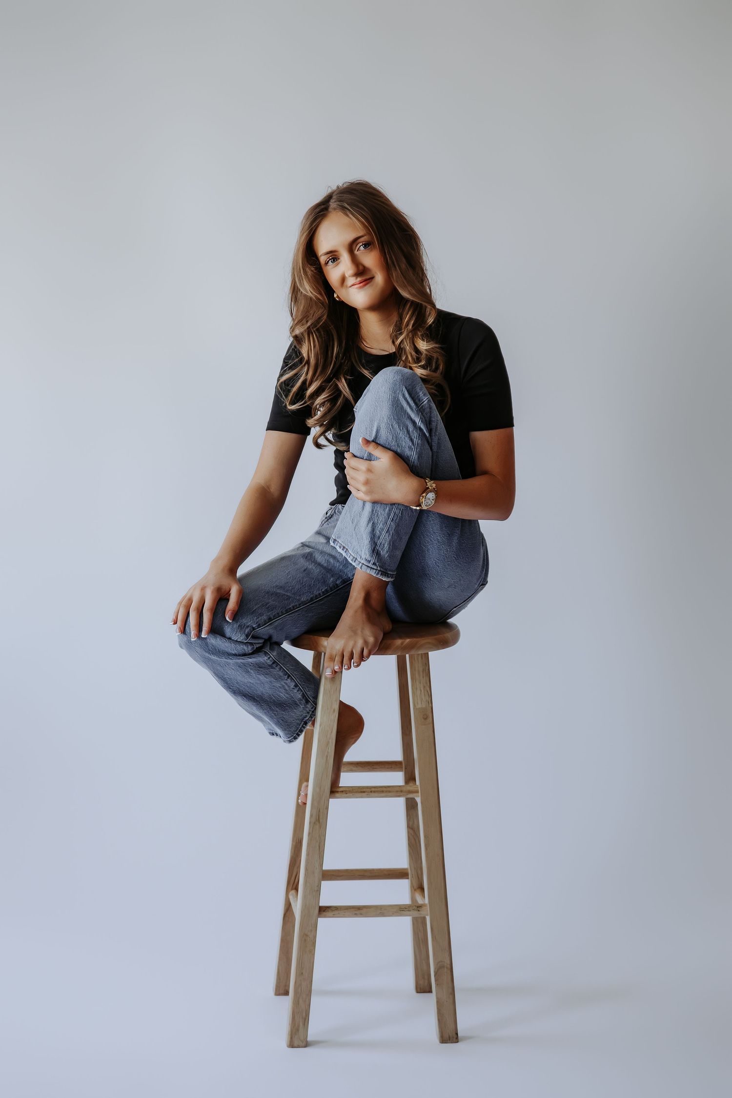 Young woman sitting on wooden stool, wearing black tee and jeans, hugging knees, smiling at camera against gray background.