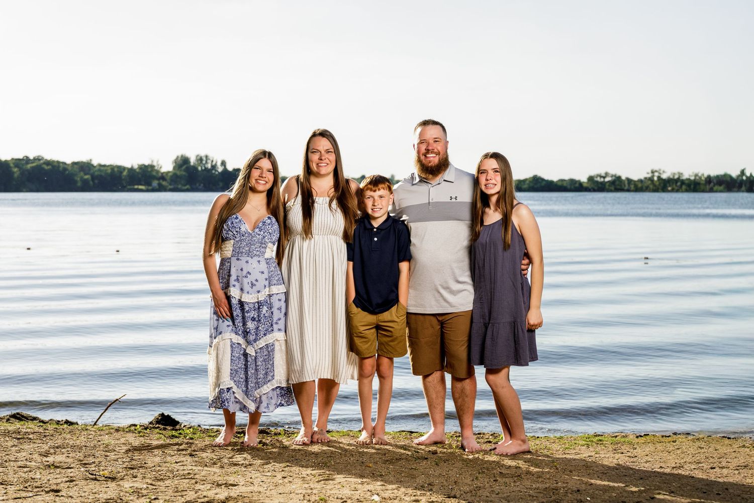 Family of five standing together on a sandy lakeside beach during golden hour sunset.