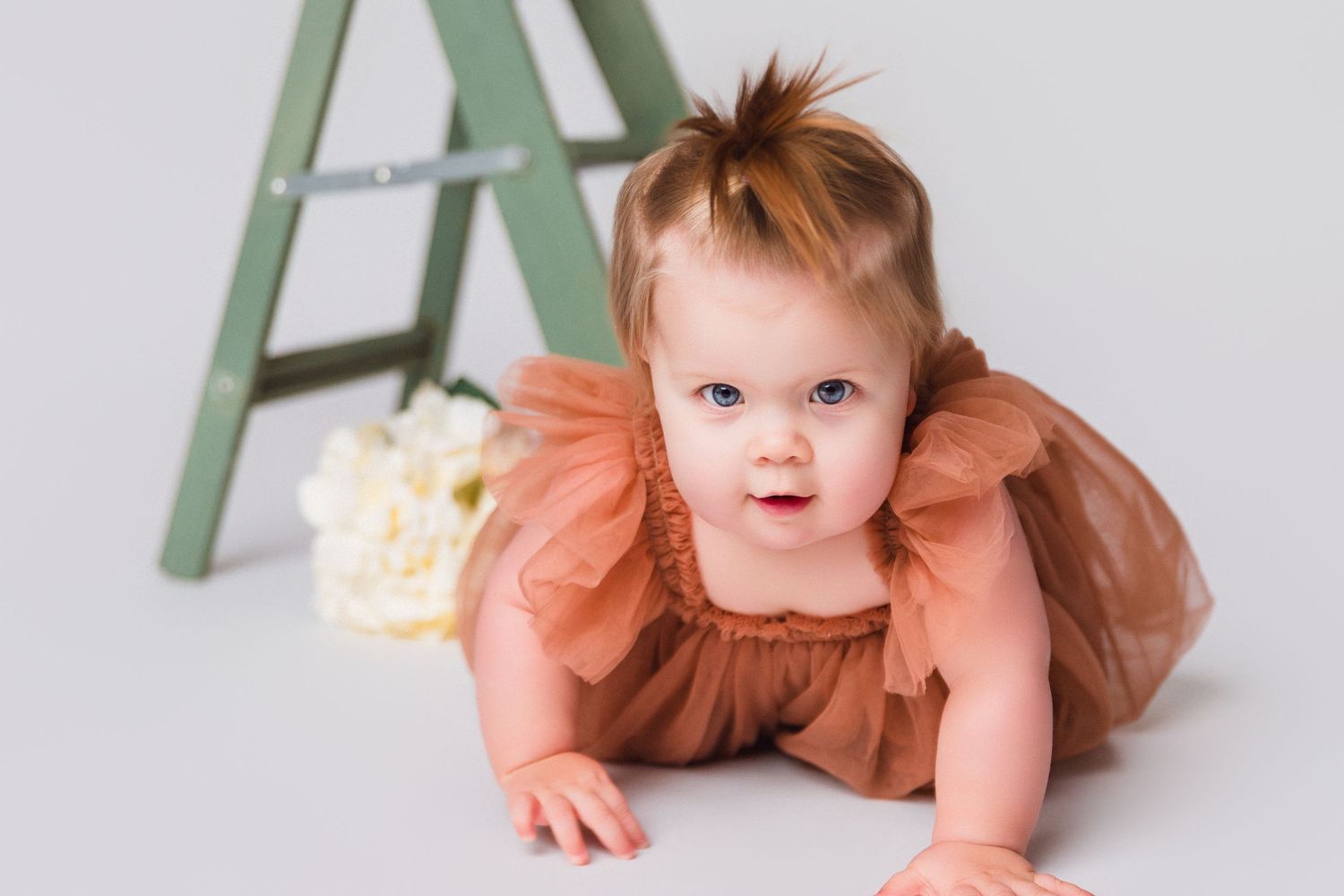 Baby in peach dress exploring near wooden ladder setup.