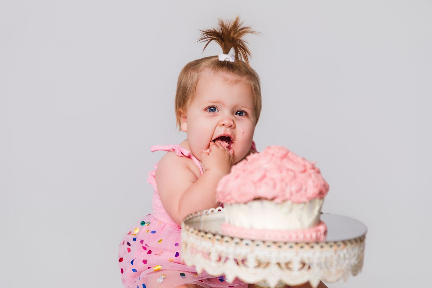 Baby in pink dress reaching for white frosted cake on ornate stand during cake smash photo session.