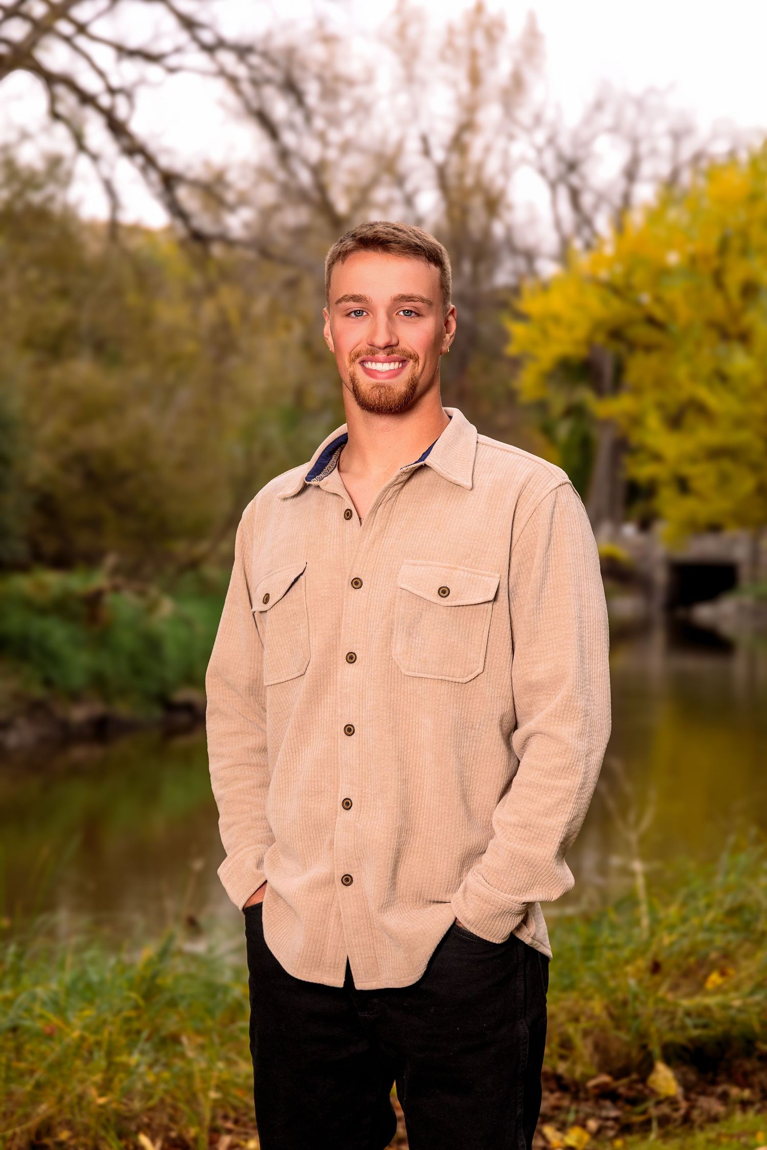 Person in beige button-up shirt smiling while standing outdoors in autumn setting.