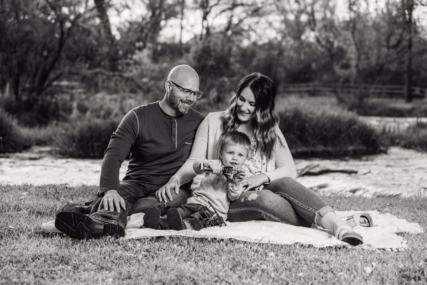 Black and white family portrait of parents and young child sitting on blanket outdoors in park setting.