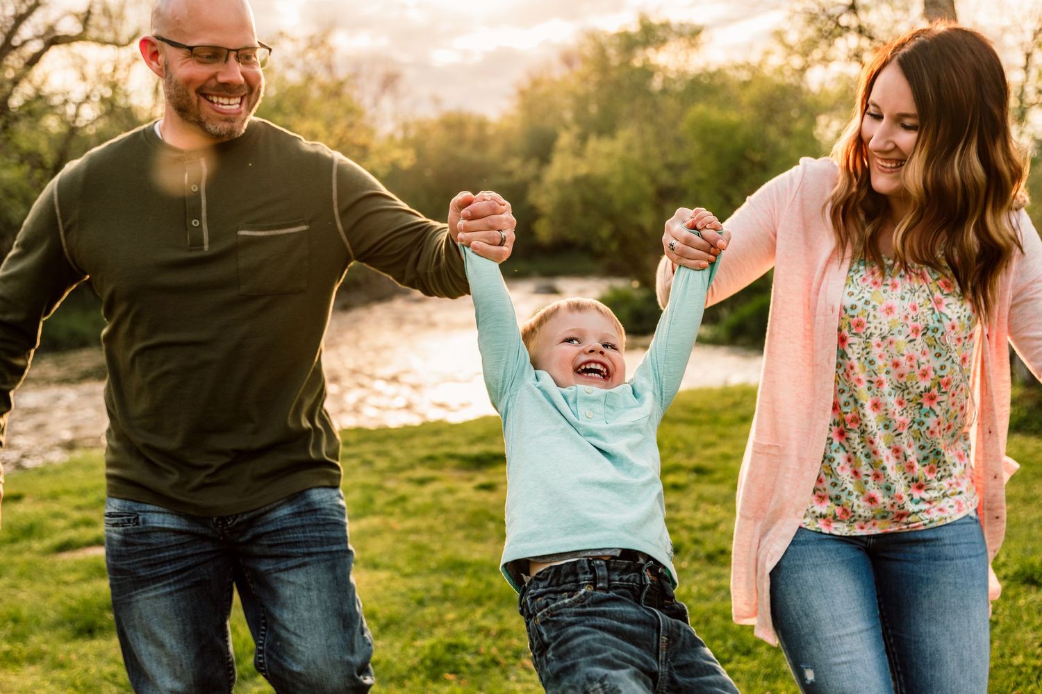 Happy family with parents swinging laughing toddler by hands outdoors near a river in a park.
