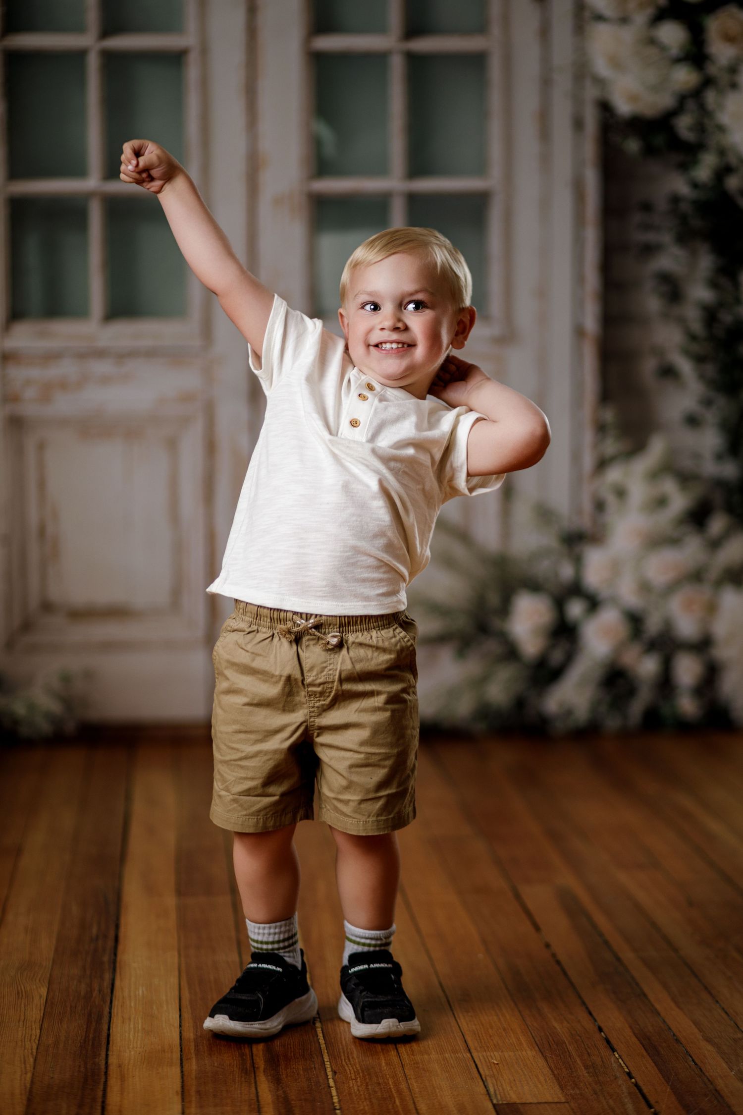 Smiling toddler boy in white shirt and khaki shorts raises fist triumphantly on hardwood floor near floral backdrop.