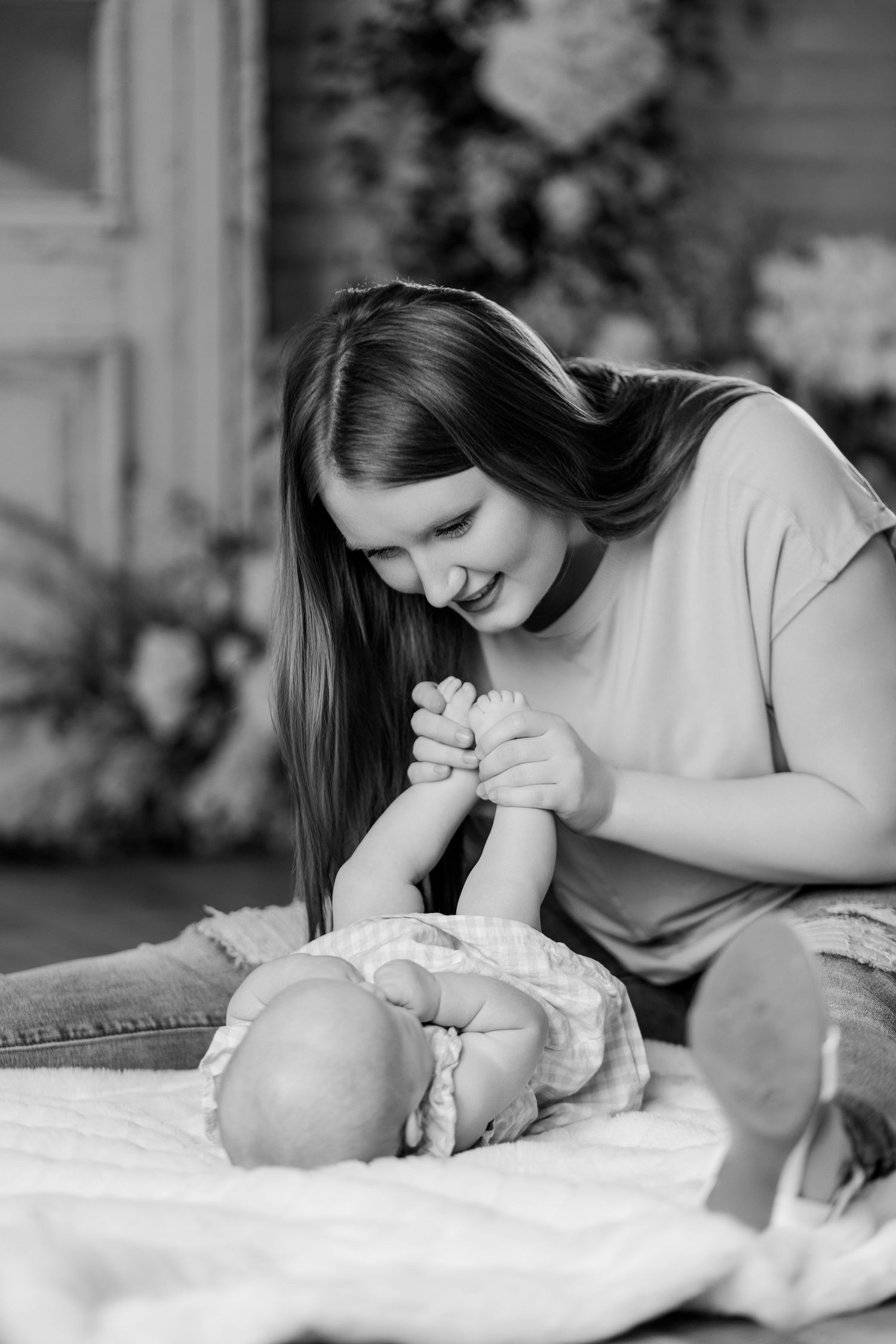 A smiling mother tenderly holds her newborn baby's hands in a black and white portrait photo.