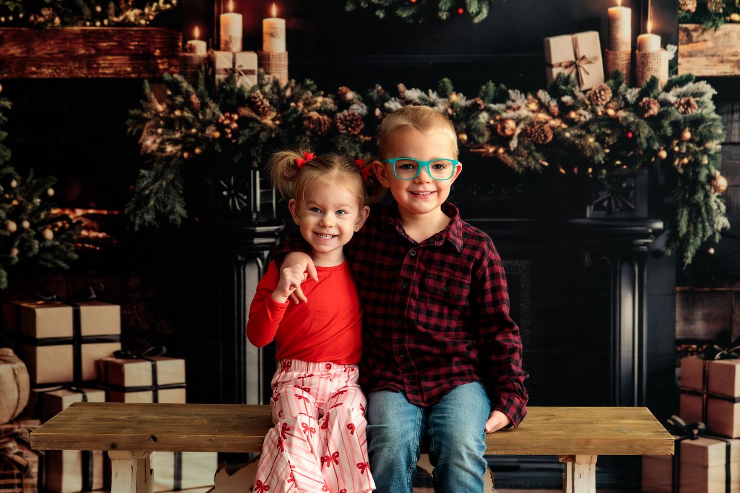 Two children sit together on a wooden bench in front of a decorated holiday mantel with candles and garland.