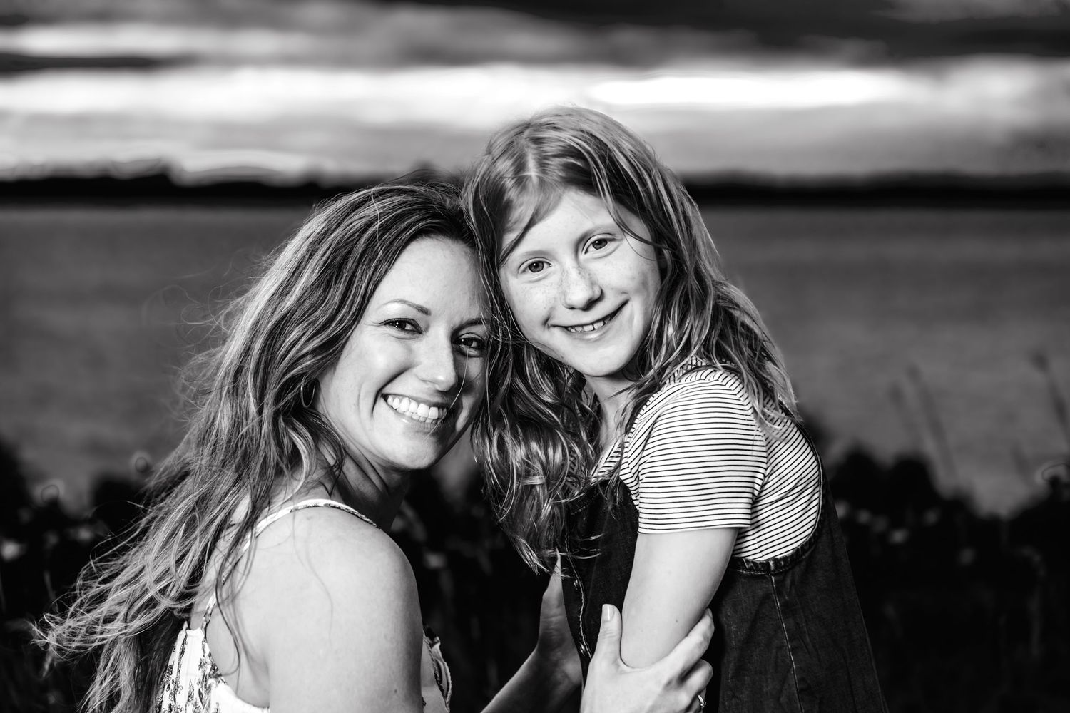Black and white portrait of two women with long hair outdoors at sunset smiling while posing together.
