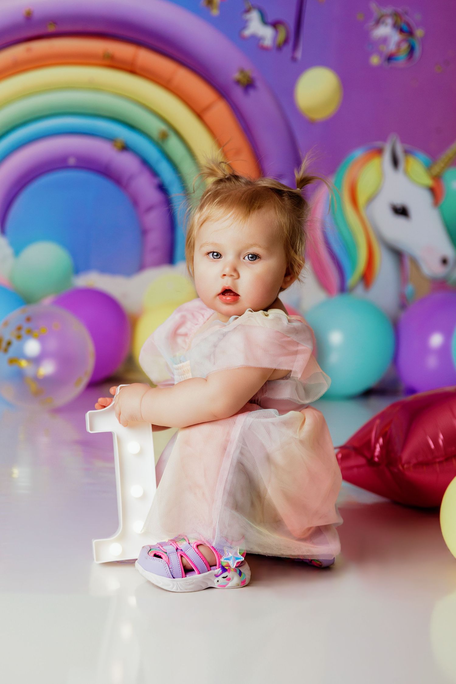 Baby girl in pink dress sits on number 1 with unicorn rainbow balloon backdrop.