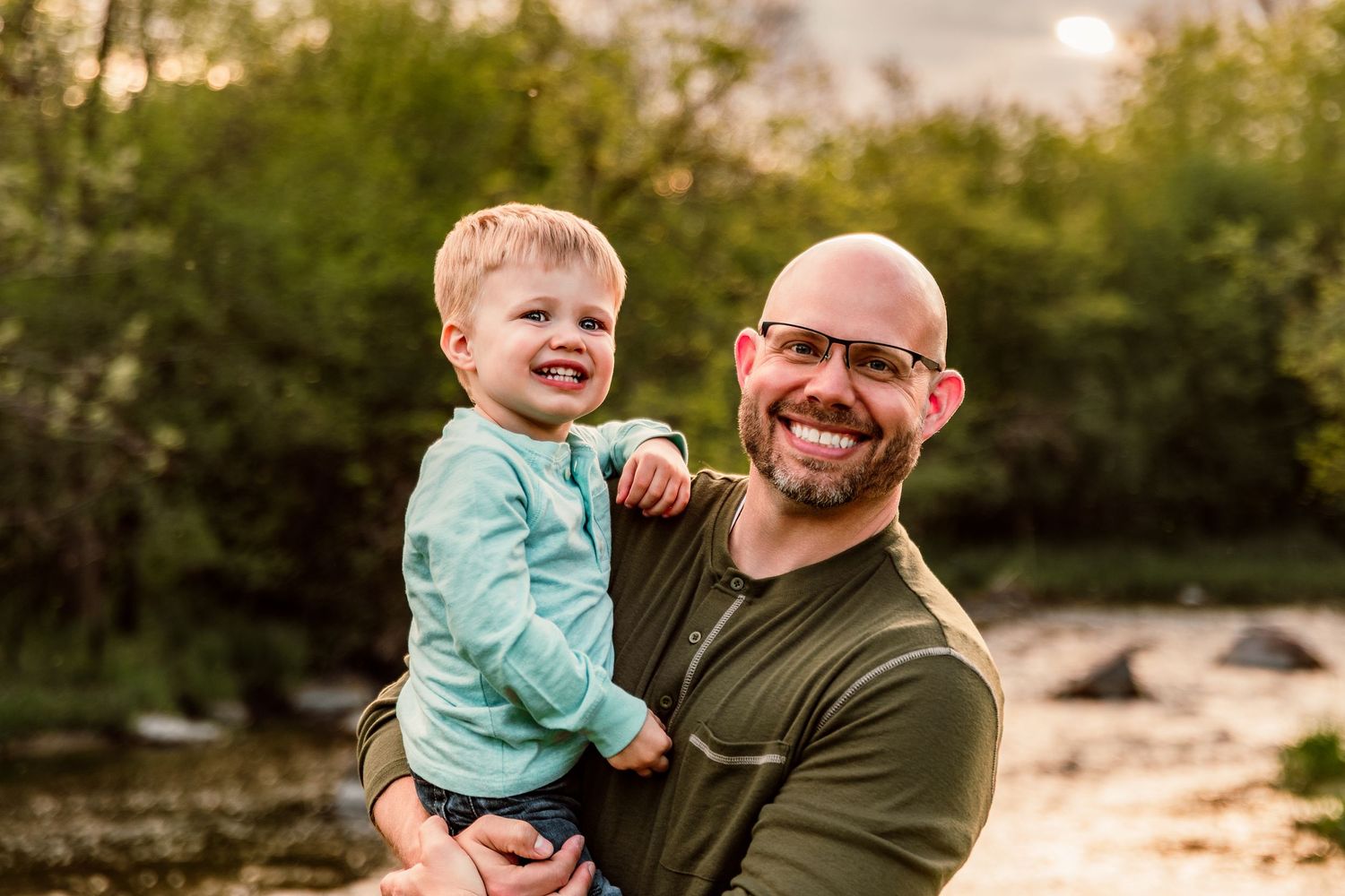 Smiling bald father with glasses holds laughing toddler boy outdoors near a creek with green trees at golden hour.