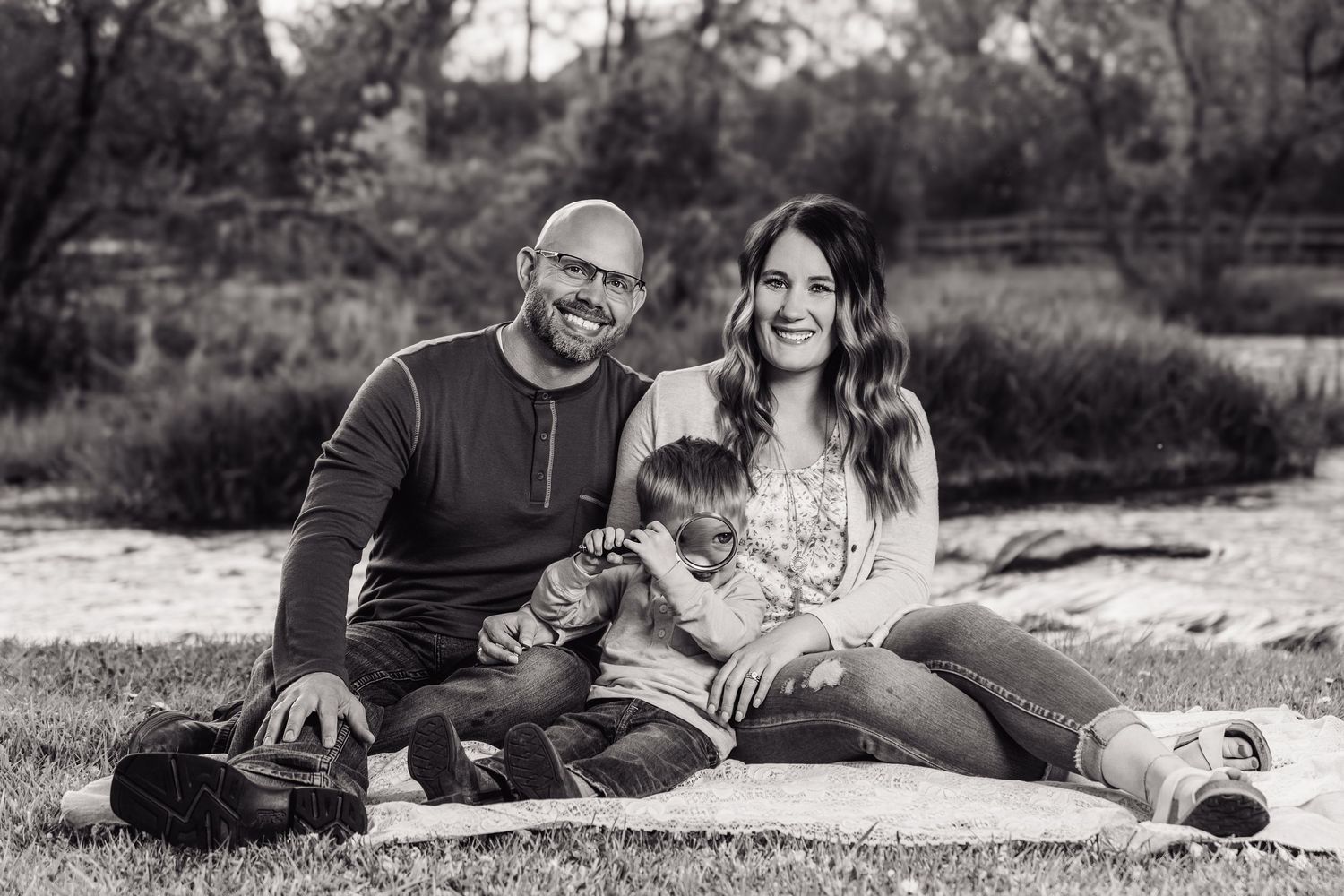 Black and white family portrait of a bald man, woman, and young child sitting together on grass outdoors.