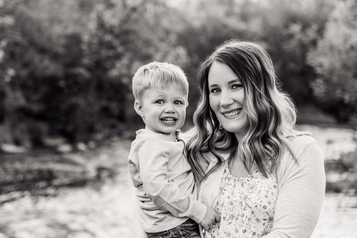 Black and white photo of a smiling mother holding her happy toddler son outdoors near trees.