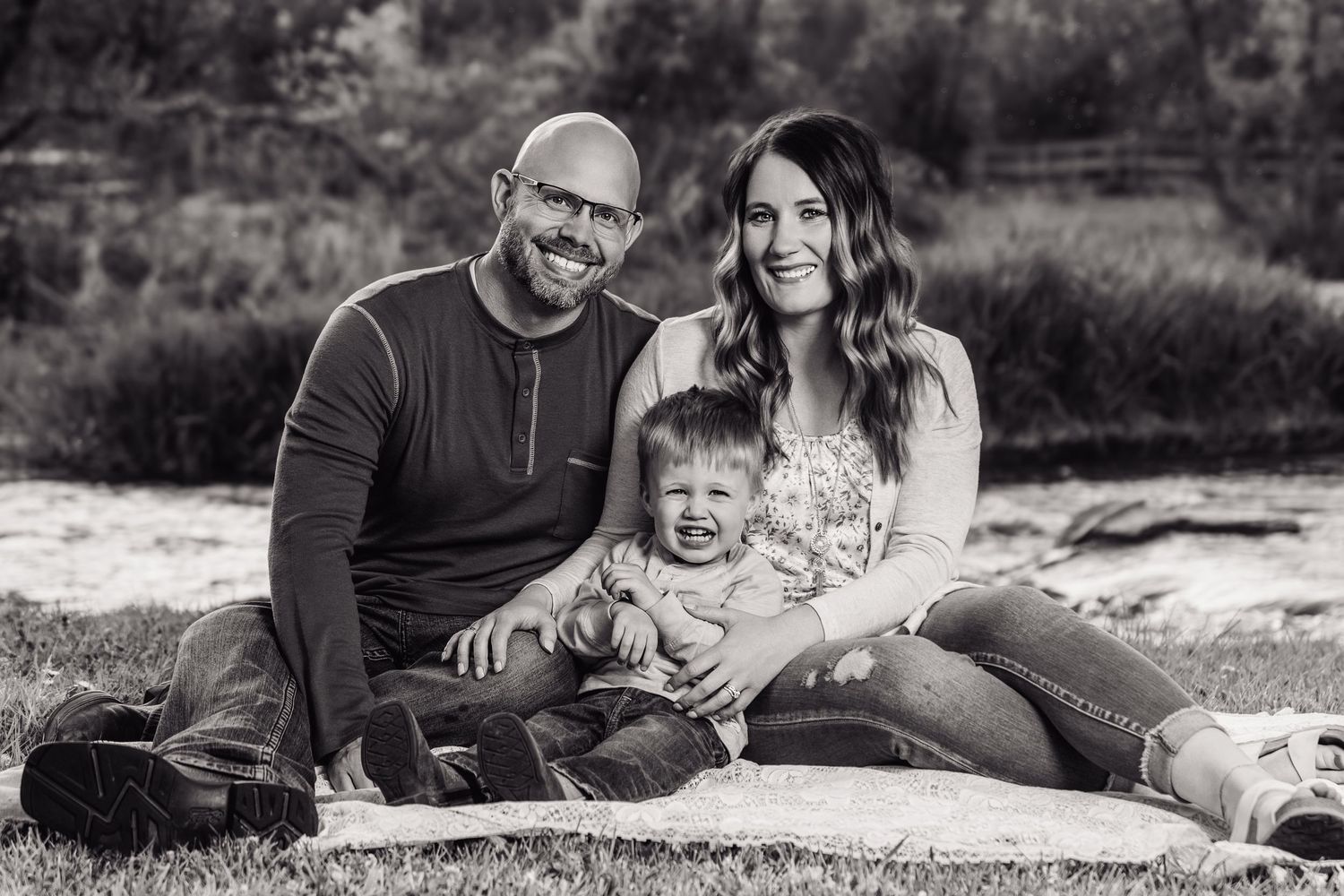 Black and white family portrait of smiling couple with toddler sitting on blanket outdoors near trees.