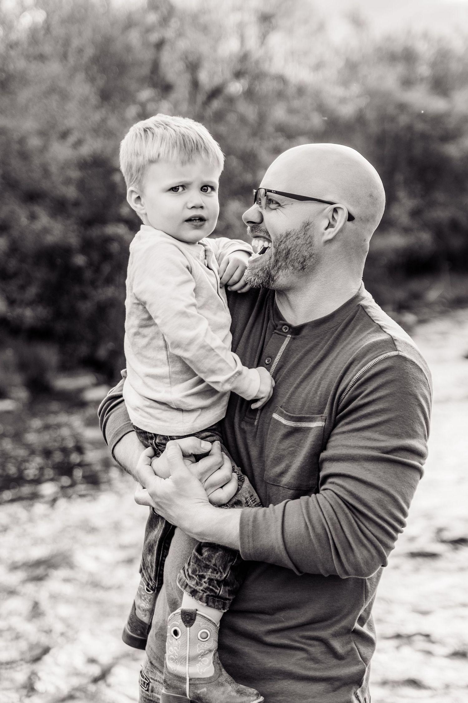 Black and white photo of a bald bearded father holding his young toddler son outdoors near a creek.