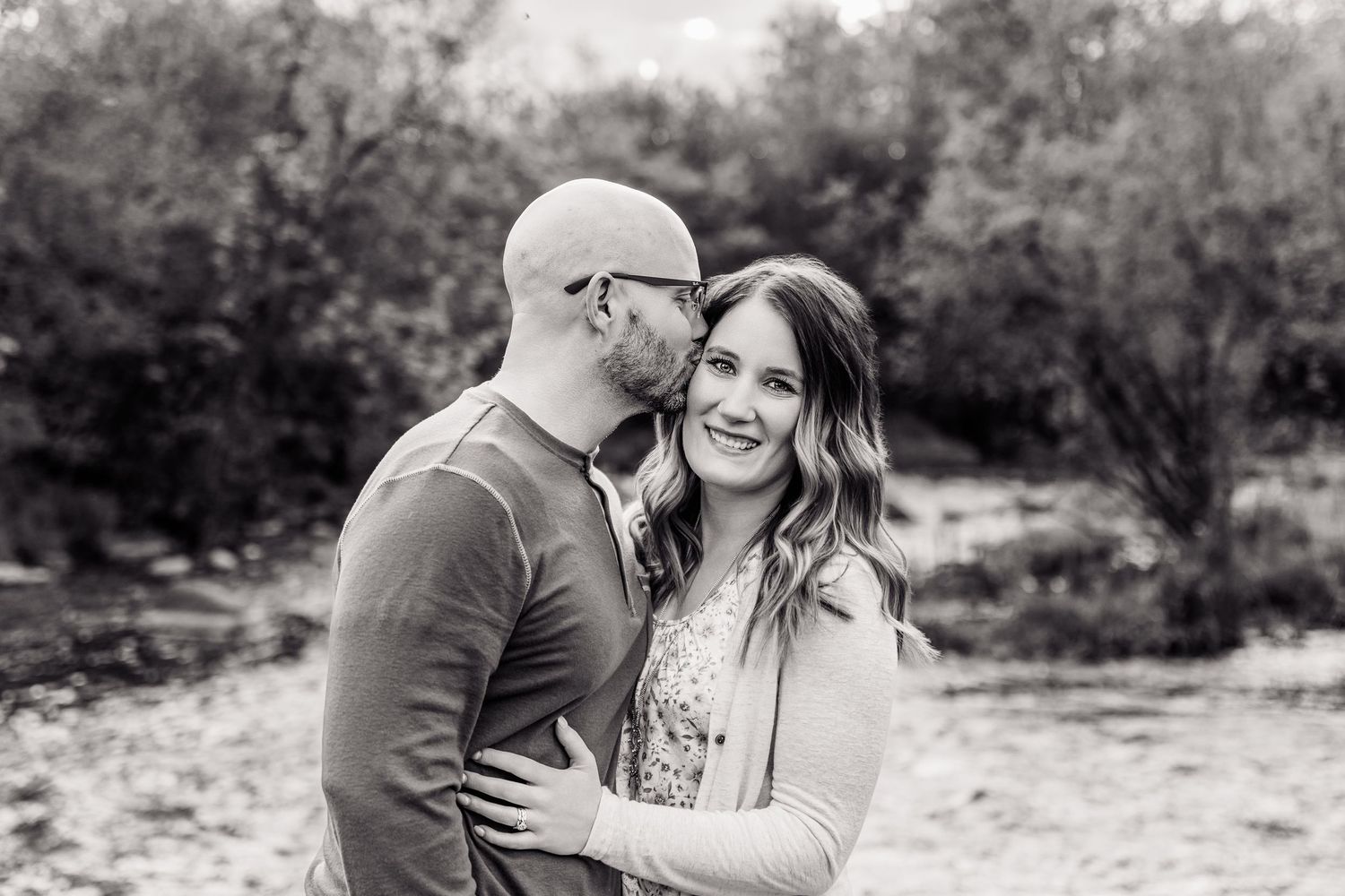 Black and white photo of a couple embracing outdoors, man kissing woman's cheek as she smiles warmly.