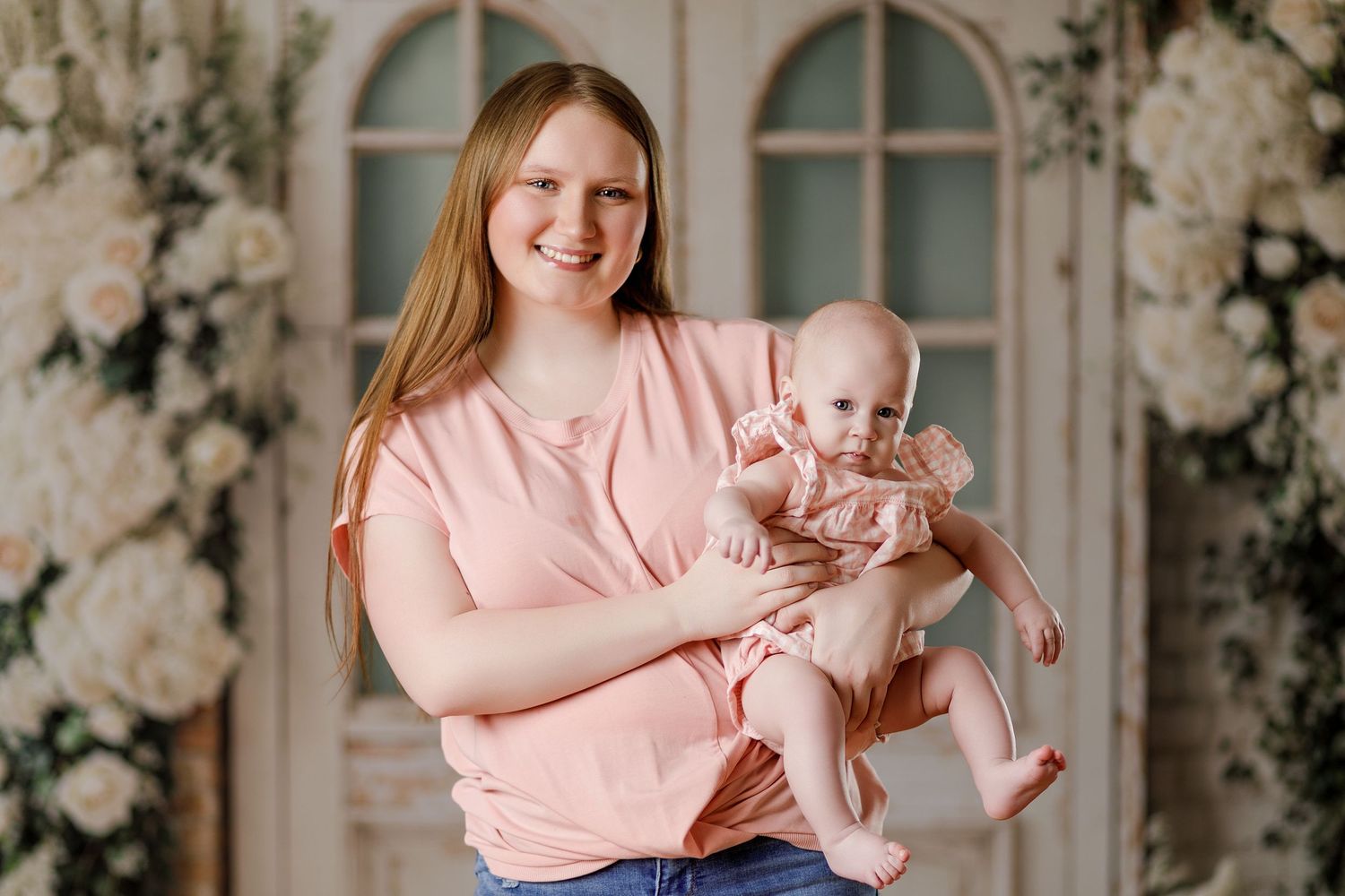 Young mother in pink top holds baby girl in pink dress, smiling in floral studio setting.