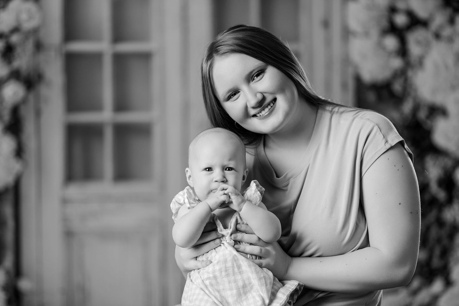 Black and white photo of smiling mother holding baby in checkered outfit in studio setting.