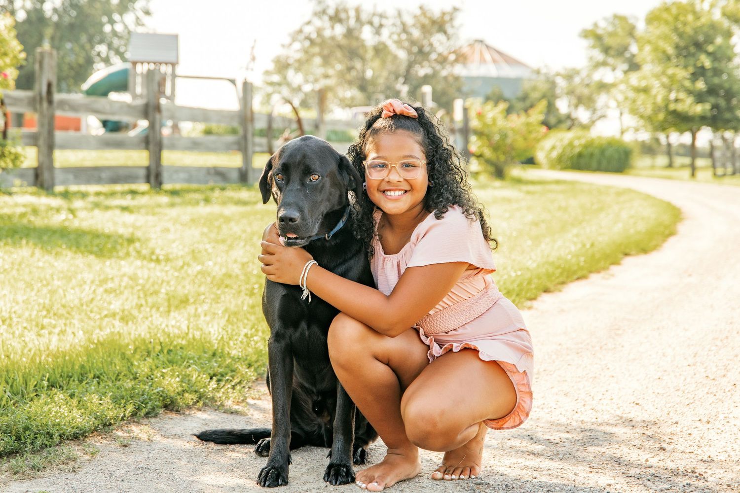 Person in pink outfit hugging a black Labrador dog on a sunny path outdoors.