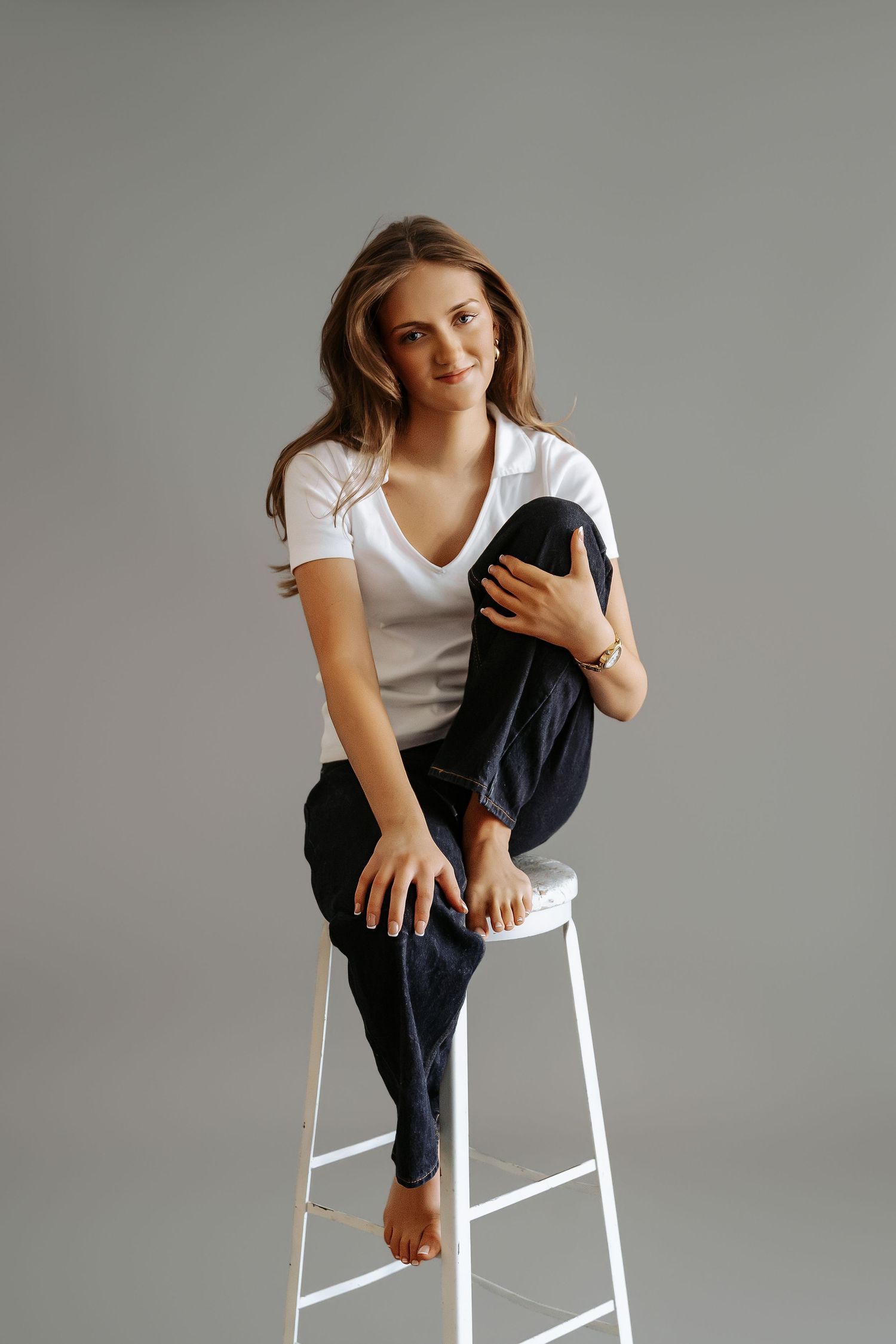Smiling young woman sitting on white stool hugging knees, white t-shirt and dark jeans.
