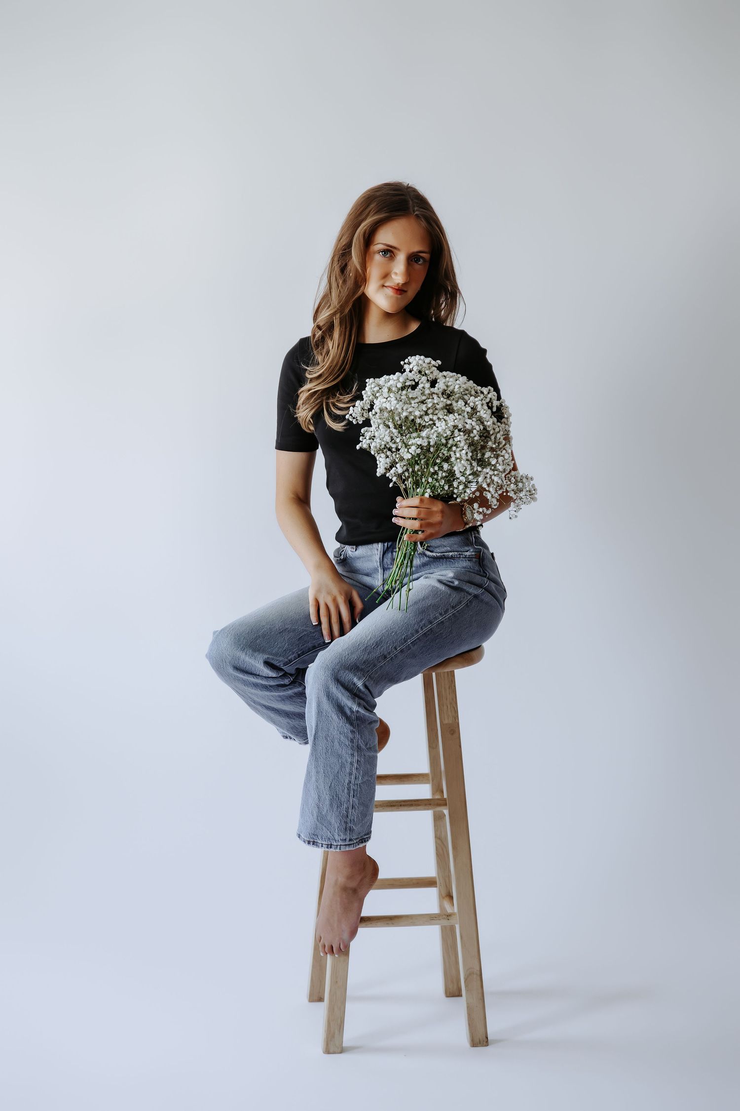 Young woman sitting on wooden stool holding white baby's breath flowers, wearing black tee and jeans, barefoot, gray background.